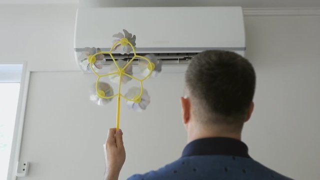 An adult man standing in front of the air conditioner holding a children's toy with a rotating propeller.