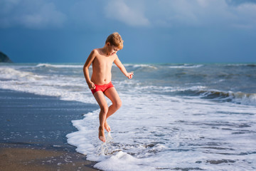 Fototapeta premium Blonde boy running and jumping on the beach on blue sea shore in summer vacation at the day time. Blue ocean with white big wawes on the background