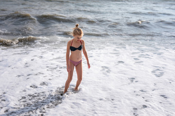 Adorable happy blonde girl in the white, red and blue swimming suit on beach vacation. Blue sea with white waves on the background