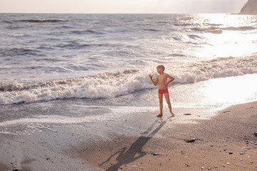 Blonde boy running and jumping on the beach on blue sea shore in summer vacation at the day time. Blue ocean with white big wawes on the background