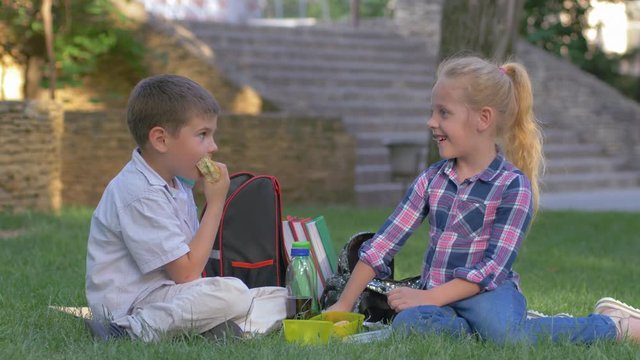 schoolchildren chat during recess lunch with sandwiches in their hands sitting on grass in schoolyard
