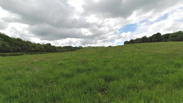Fast motion clouds over a large grass covered field surrounded by woodland.