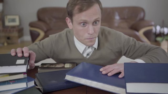 Young modestly dressed man fell asleep at the desk in the office, many books are on the table and on his head. The frustrated and tired nerd waking up and looking around. The guy study too much