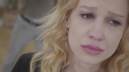 Close up blond woman in the foreground crying, an emotional angry man yelling and shouting on the background. The couple has a quarrel. Difficulties in relationship