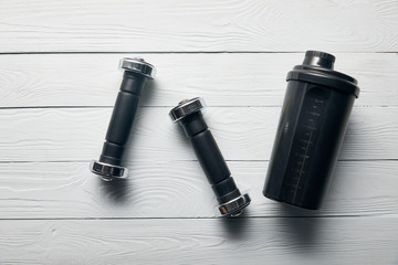 top view of black shaker cup and dumbbells on wooden white background