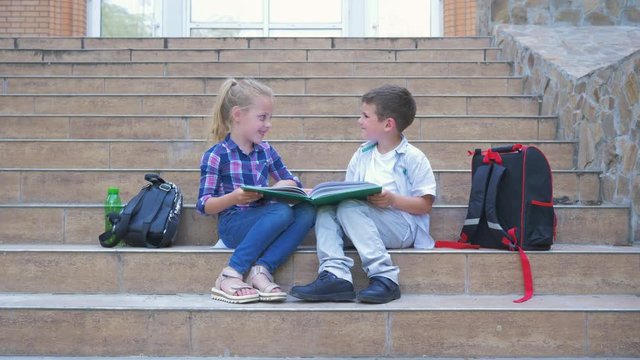 school, little classmates with backpacks sitting on the steps and view pictures in a book during break outdoors