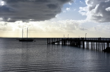 Fototapeta na wymiar Kingfisher Bay pier at sunset. Moored sailboats and yachts in the bay. Silhoutette of people on the pier. World Heritage-listed Fraser Island is the world’s largest sand island.