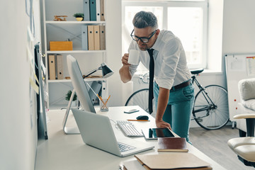 So busy. Good looking young man drinking coffee and smiling while standing in the office