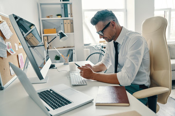 Texting to client. Thoughtful young man in shirt and tie using smart phone while sitting in the office