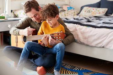 Single Father At Home With Son Teaching Him To Play Acoustic Guitar In Bedroom