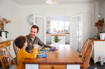 Single Father Sitting At Table Eating Meal With Son In Kitchen At Home