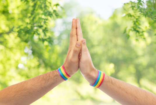 lgbt, same-sex love and homosexual relationships concept - close up of male couple hands with gay pride rainbow awareness wristbands making high five gesture over green natural background