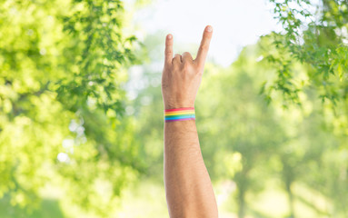 lgbt, same-sex relationships and homosexual concept - close up of male hand wearing gay pride awareness wristband showing rock or hand-horns sign over green natural background