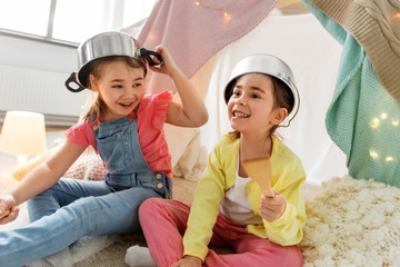 childhood, hygge and friendship concept - happy girls with cooking pots playing in kids tent at home