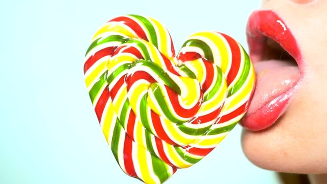 close-up, female lips with red lipstick, eat and lick a big lollipop in the shape of a heart. copy space, blue background