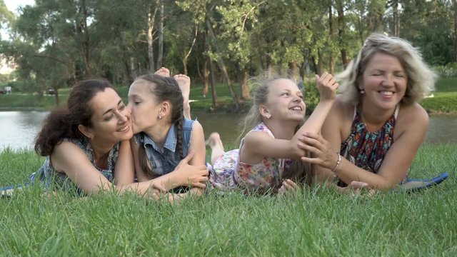 Family of the same sex: mothers with young daughters have fun and play lying on the grass in the Park at the sunset of a summer day by the river. A family holiday in the fresh air. Concept. 4K