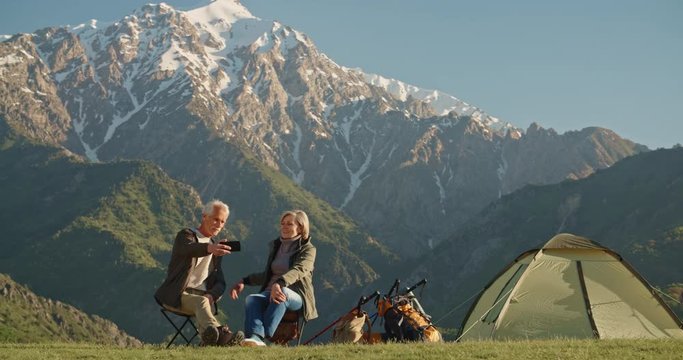 Senior cacasian couple having a rest on top of a mountain, taking a picture or having video chat on smartphone, travelling together after retirement - pension, recreational pursuit, tourism concept 4k