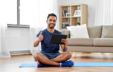 sport, technology and healthy lifestyle concept - smiling indian man with tablet computer sitting on exercise mat and showing thumbs up at home