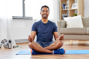 fitness, meditation and healthy lifestyle concept - indian man meditating in lotus pose on exercise mat at home