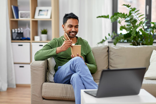 Consumption And People Concept - Smiling Indian Man Eating Takeaway Food And Watching Something On Laptop Computer At Home