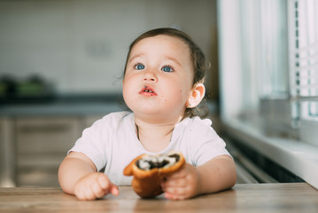 a little girl in the kitchen during the day eating a bun with poppy seeds is very appetizing