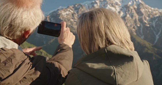 Senior cacasian couple having a rest on top of a mountain, taking a picture on smartphone, travelling together after retirement - pension, recreational pursuit, tourism concept 4k