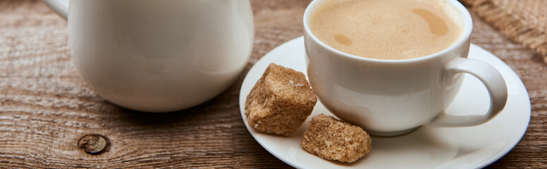 panoramic shot of delicious coffee with foam in cup on saucer with brown sugar near sackcloth and milk jug on wooden background