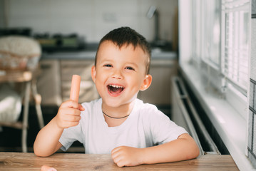 Portrait of a adorable little boy in a white t-shirt at the table eating a sausage with his hands