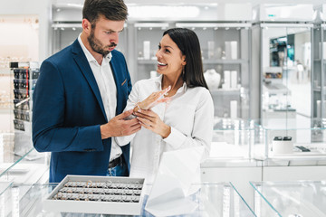 Beautiful couple enjoying in shopping at modern jewelry store. Young woman try it out gorgeous...
