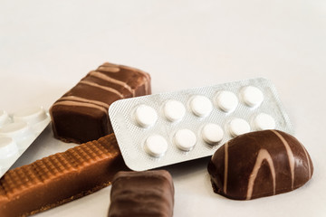 Chocolate candies and pills lie on a white table. Concept of dangerous neighborhood medicaments and sweets. Close-up. Limited depth offield.