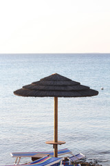 Straw umbrella against blue sea and sky. Two deckchairs remain under the umbrella.
