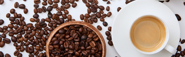 top view of coffee in cup on saucer near scattered roasted beans and wooden bowl, panoramic shot