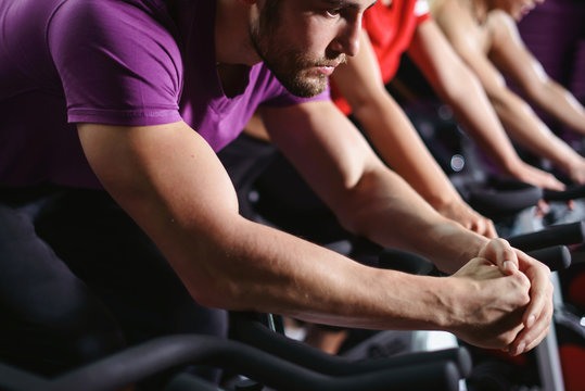 Close up hands of man biking in spinning class. Group of smiling friends at gym exercising on stationary bike. Happy cheerful athletes training on exercise bike.