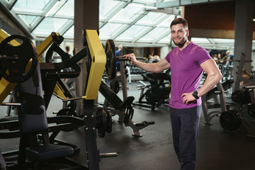 Fototapeta na wymiar Handsome friendly personal trainer in gym. Young sporty man smiling standing on fitness club background