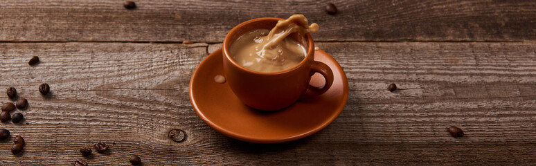 panoramic shot of coffee splash in cup on wooden table with scattered coffee beans