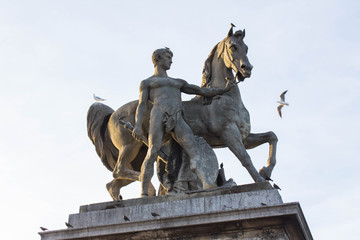 Obraz premium The statue near the bridge of a nude man with a sword in one hand and holding a horse by the bridles. Bright summer day and seagulls on a monument. Art and architecture in the Paris.
