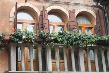 Fototapeta na wymiar Balcony with potted flowers and antique shutters on the windows on the facade of a residential building in the courtyard of Venice in Italy. Three windows on the balcony with columns.