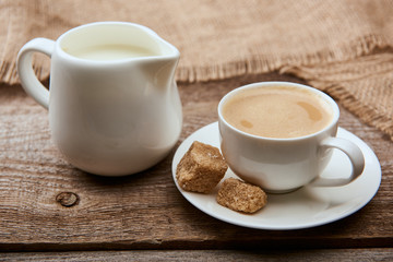 delicious coffee with foam in cup on saucer with brown sugar near sackcloth and jug milk on wooden background