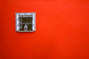 Red big space of scarlet wall of the building and small white square window on it. Window with black wiggling grid and small metal lamp on the window sill. Copy space for designers. Residential area.