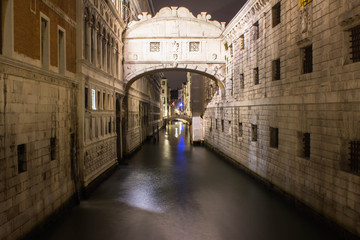 Obraz na płótnie Canvas Venice's wide canal at night, night lights reflecting in the water. An ancient arch over the canal connecting two buildings. Arched bridge in the background. Old traditional architecture at night.