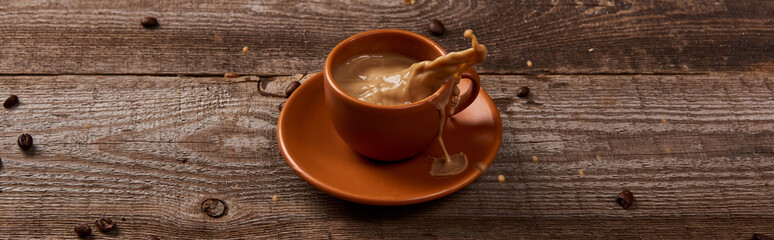 panoramic shot of coffee splash in brown cup on wooden table with scattered coffee beans