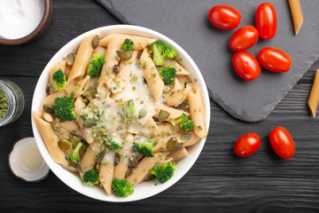 Bowl with pasta, green onions, white sauce, pumpkin seeds and broccoli. On the table are cherry tomatoes, salt, green onions. 