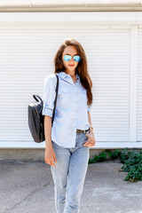 Young stylish woman wearing blue shirt, blue denim jeans, black backpack and sunglasses posing outdoors against white street wall. Trendy casual spring summer outfit. Street fashion.