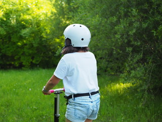 Girl on a scooter seen from behind in park, wearing a white helmet and denim shorts