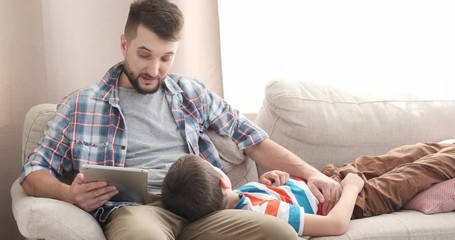 Father reading ebook on digital tablet to his son relaxing on sofa at home - Powered by Adobe