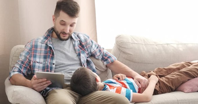 Father reading ebook on digital tablet to his son relaxing on sofa at home