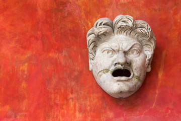 Antique mask from a Roman comedy: unpleasant frowning face with open mouth. Red background and copy space.