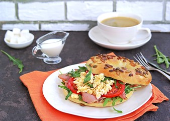 Breakfast, bagel with scramble egg, ham, arugula and vegetables on a white plate on a dark background.