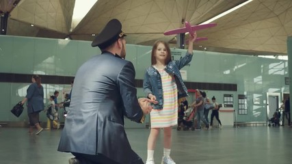 Joyous smiling pilot presenting toy pink plane to a little cute girl in terminal of airport.