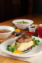Close up of a roasted pork cutlet with boiled bulgur and greens as a part of set meal in a restaurant, with other meals on a background